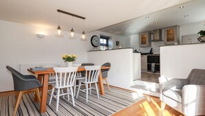 A modern kitchen with white cabinets, a black clock, and a dining area with a striped rug and wooden table. - Great Meadow Barn (Newquay)