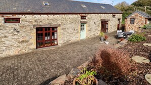 A stone building with a paved courtyard, a small garden with plants, and a seating area with outdoor furniture. - Great Meadow Barn (Newquay)