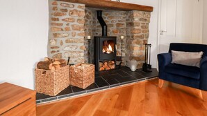 A cozy living room with a stone fireplace, a blue armchair, and wicker baskets filled with firewood. - Great Meadow Barn (Newquay)