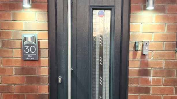 A black door with a vertical handle, a glass panel, and a metal mail slot. There are two wall-mounted light fixtures on either side of the door. A sign on the left reads "30 The Annex."
