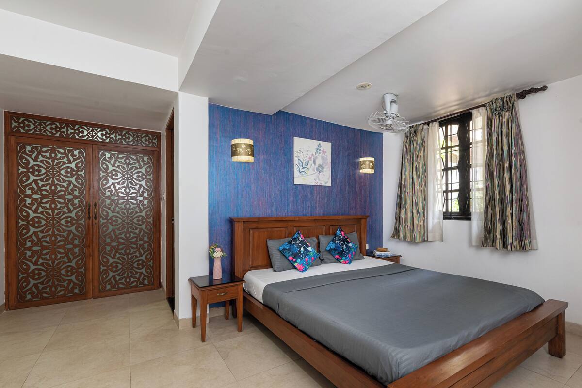 A bedroom with a wooden bed, a nightstand, a wall-mounted lamp, and a window with curtains.
