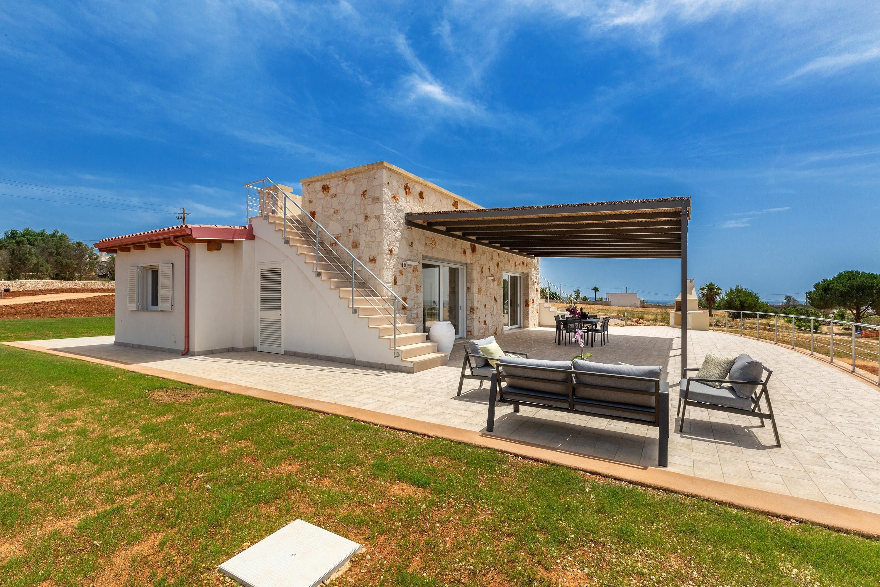 A modern house with a stone exterior, a covered patio with outdoor furniture, and a staircase leading to the entrance.