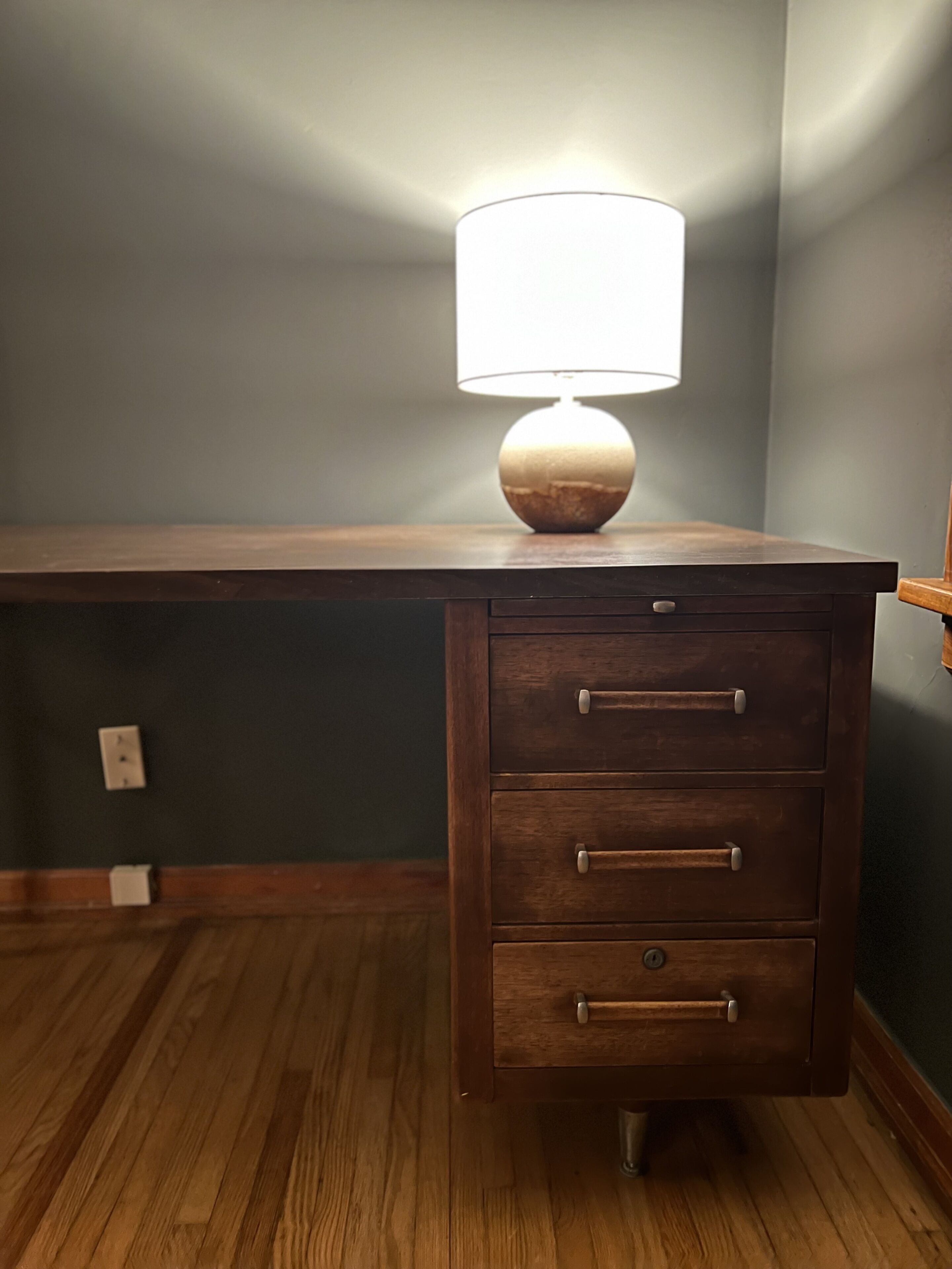 A wooden desk with a lamp, drawers, and a light switch on the wall.