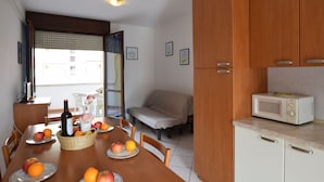 A compact kitchen with wooden cabinets, a microwave, and a dining table set with fruits and a bottle. - Lovely apartment for 6 people with pool, WIFI, A/C, TV, terrace and pets allowed (Rosolina)