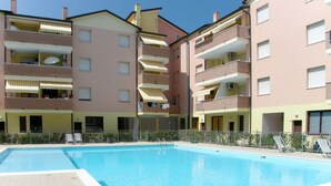 Une piscine dotée d’une terrasse carrelée, entourée d’un immeuble à plusieurs étages, agrémenté de balcons et d’auvents.