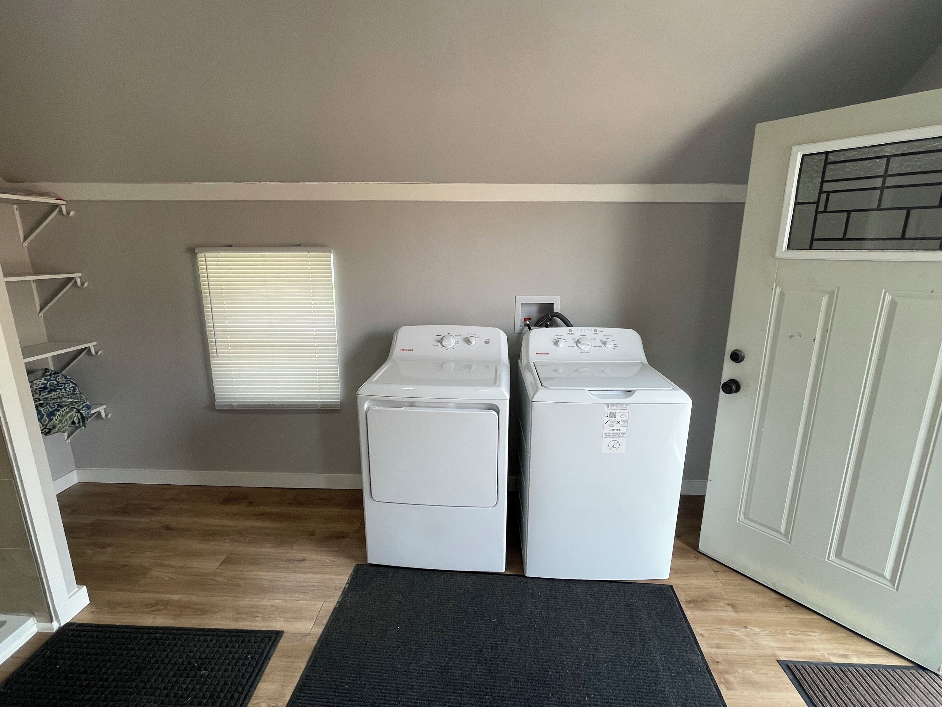A laundry room with two white washing machines, a window with blinds, and a door with a glass panel.