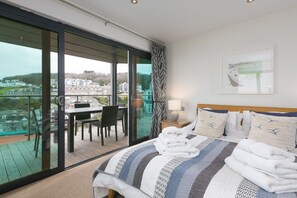 A hotel room with a large bed, a view of the city through a glass door, a dining area with a table and chairs, and a painting on the wall.