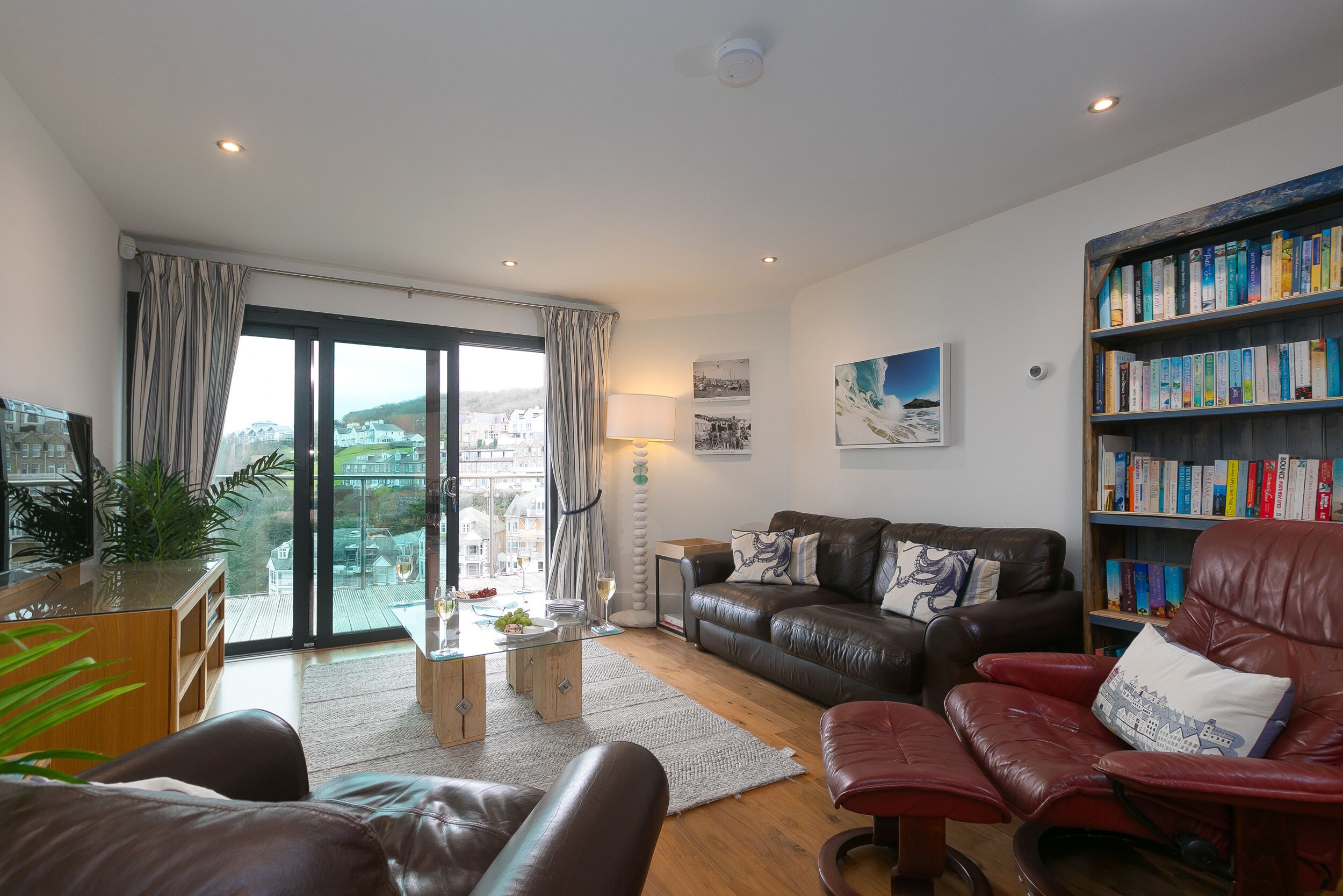 A modern living room with leather furniture, a bookshelf, and a view of the city through large windows.