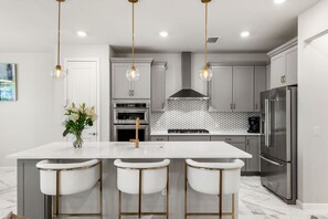 Une cuisine moderne dotée d’un îlot central, de tabourets de bar, d’appareils électroménagers en acier inoxydable et de luminaires suspendus.