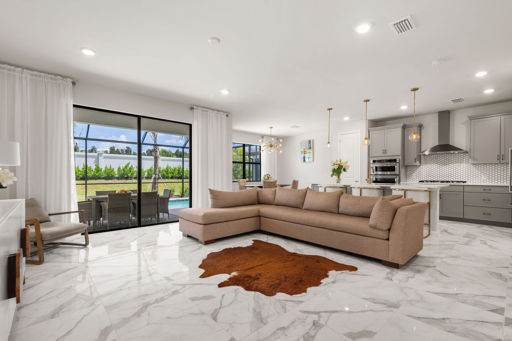 A modern living room with a sectional sofa, a glass door leading to a patio, and a kitchen with contemporary appliances and pendant lighting.