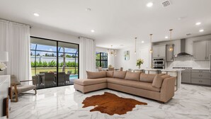 A modern living room with a sectional sofa, a glass door leading to a patio, and a kitchen with contemporary appliances and pendant lighting.