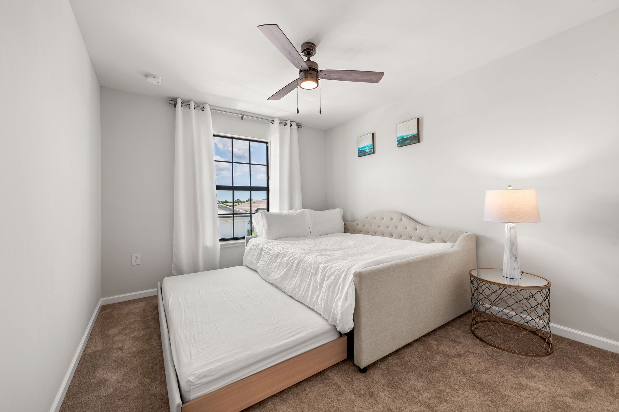 A bedroom with a bed, a side table with a lamp, a ceiling fan, and a window with curtains.