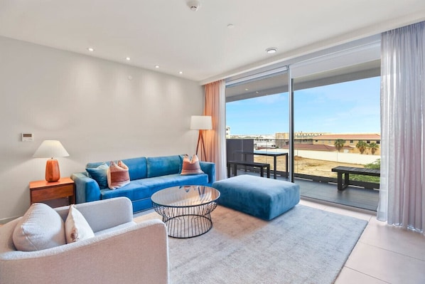 Ein modernes Wohnzimmer mit einem blauen Sofa, einem runden Couchtisch und Blick auf den Strand durch große Fenster.