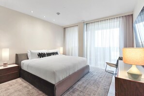 Ein modernes Hotelzimmer mit einem großen Bett, einem Nachttisch mit Lampe, einem Schreibtisch ebenfalls mit Lampe und Blick auf die Stadt durch große Fenster.