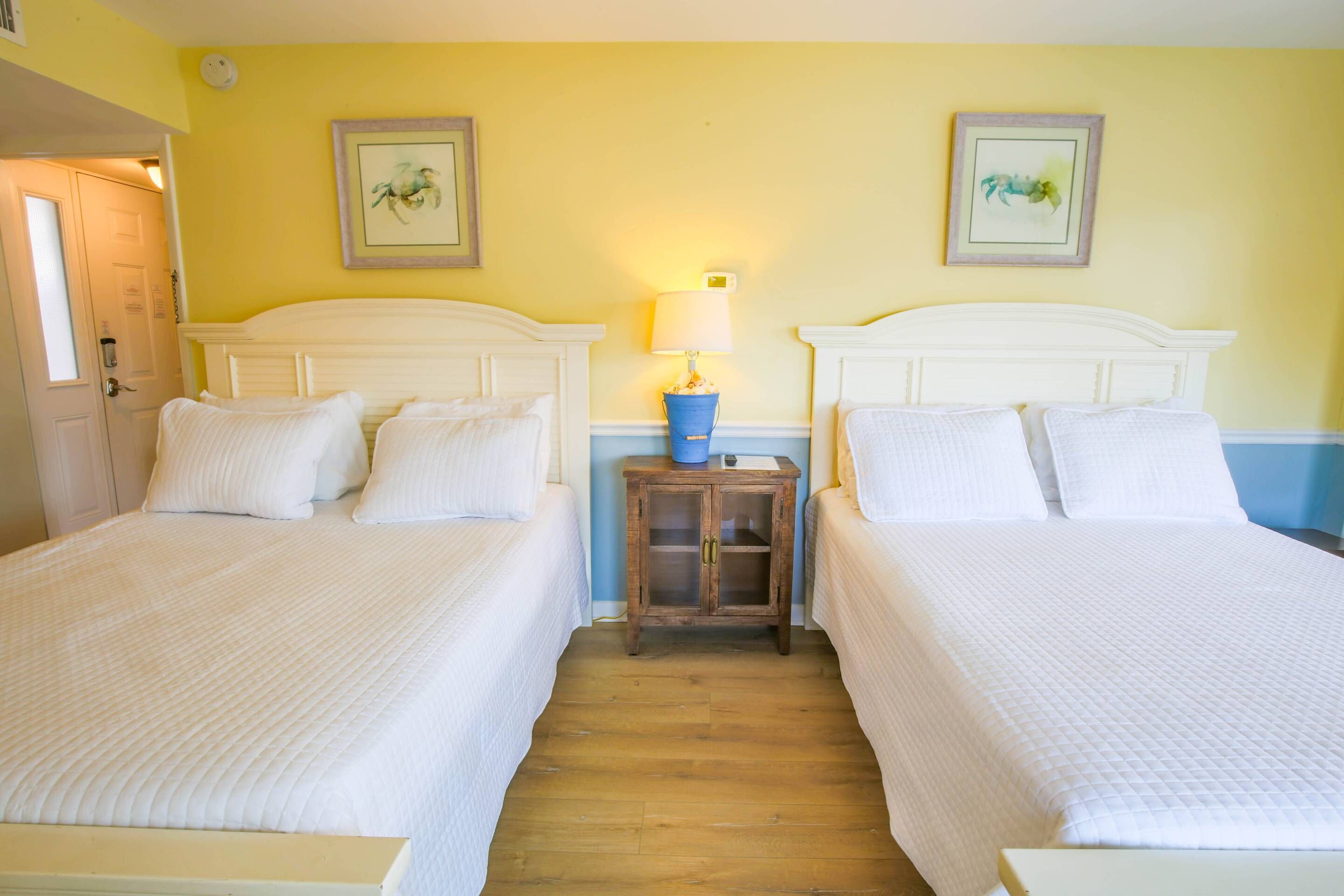Una habitación de hotel con dos camas, una mesita de noche de madera con una lámpara y dos cuadros en las paredes.