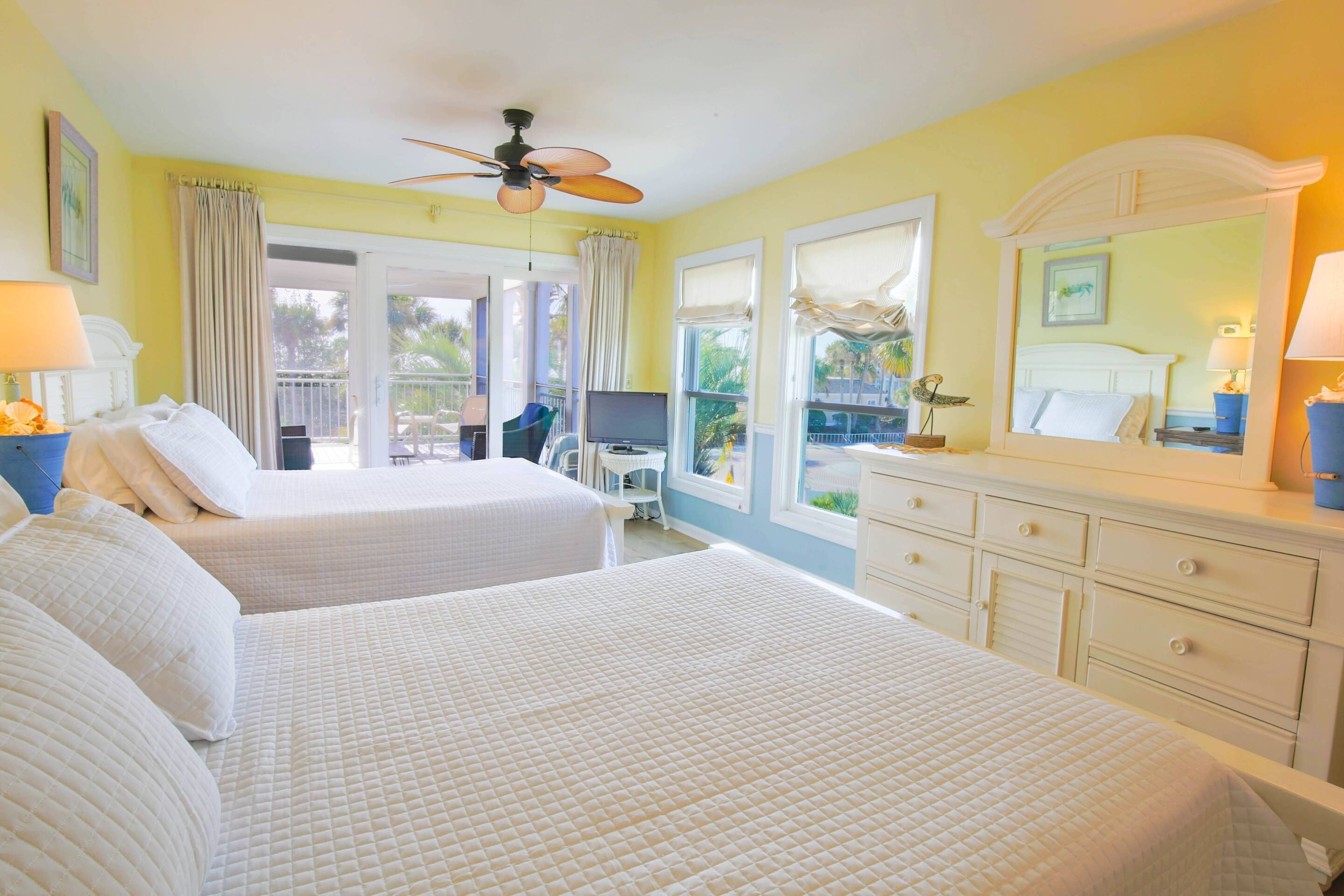 A hotel room with two beds, a ceiling fan, a TV, and a balcony with a view of the outdoors.