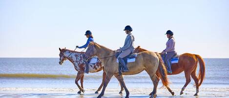 Tres personas montando caballos en una playa, con el océano al fondo.