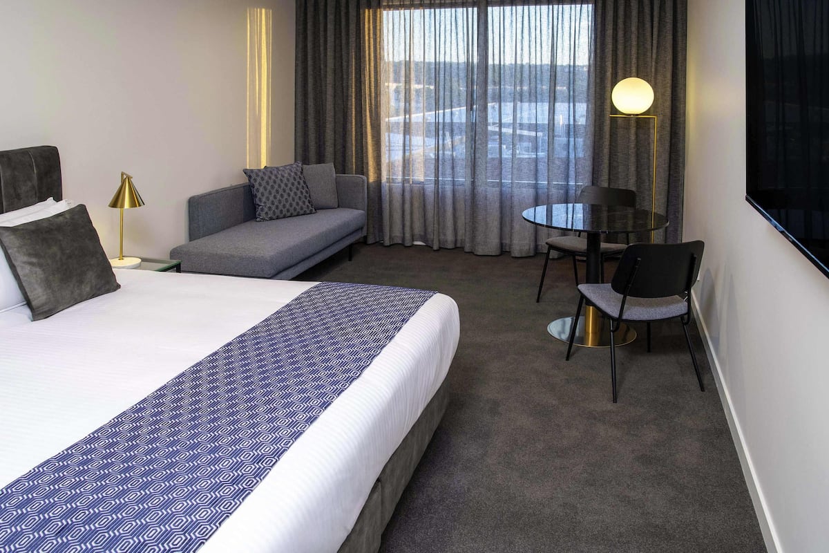 A hotel room with a bed, a sofa, a dining table with chairs, a TV, and a window with curtains.
