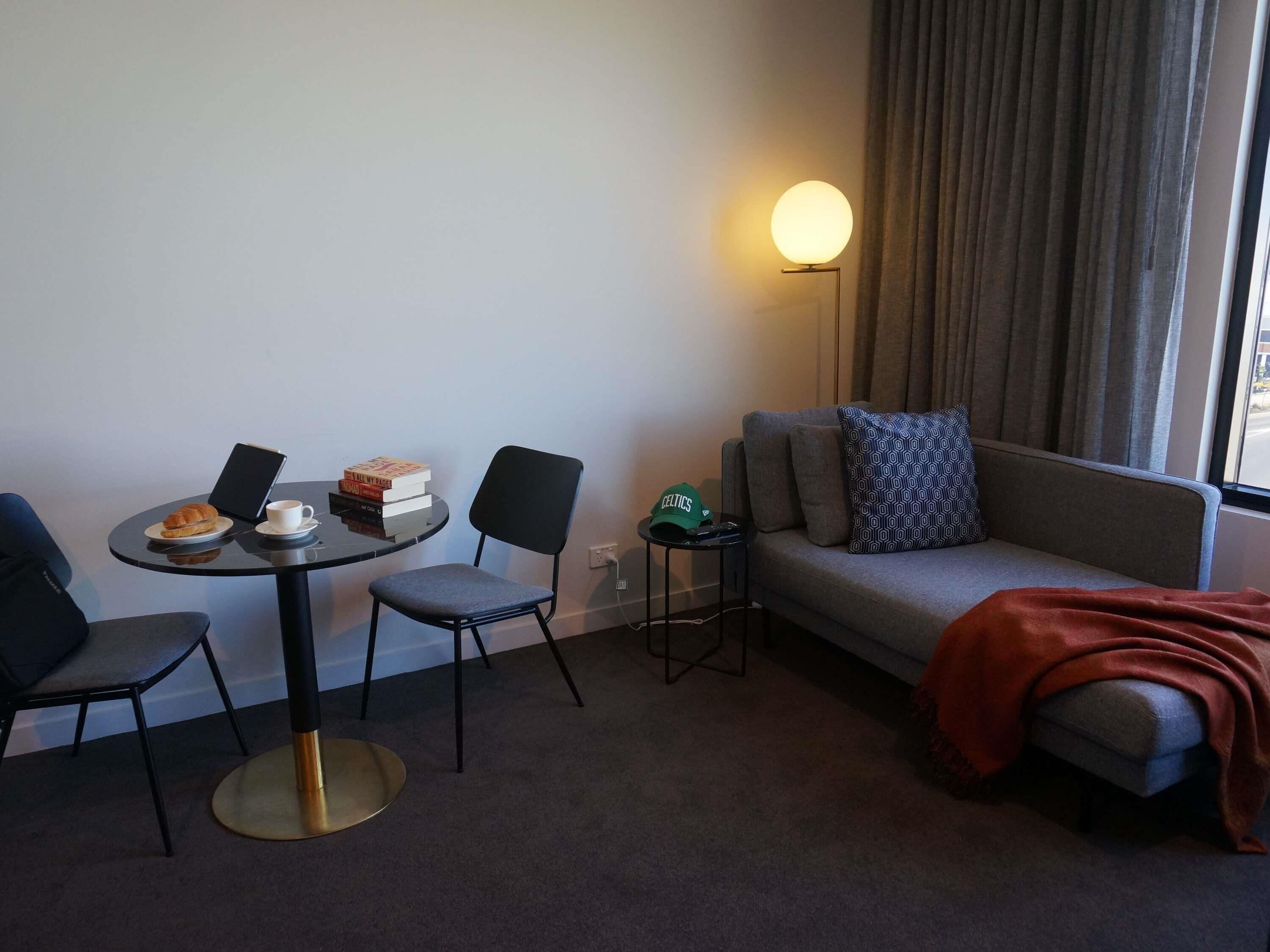Habitació d'hotel amb taula rodona, dues cadires, un sofà, una làmpada i una finestra amb cortines.