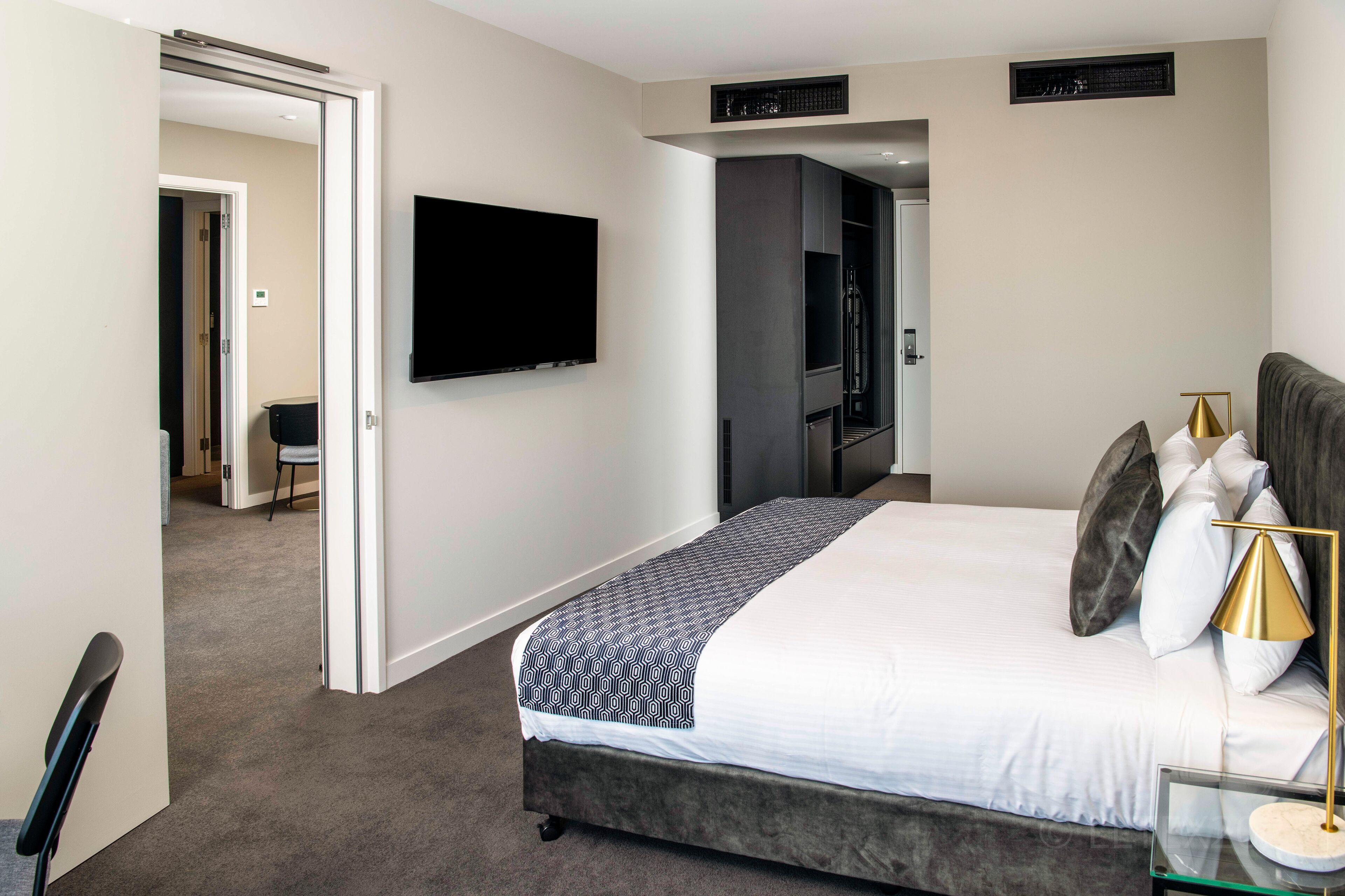 Habitación de hotel con una cama grande, un televisor de pantalla plana en la pared, una mesita de noche con lámpara y una silla en la esquina.