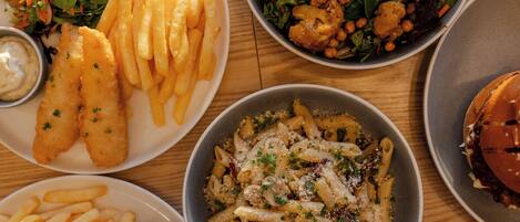 Un tavolo con diversi piatti, tra cui fish and chips, pasta, insalata e un hamburger.