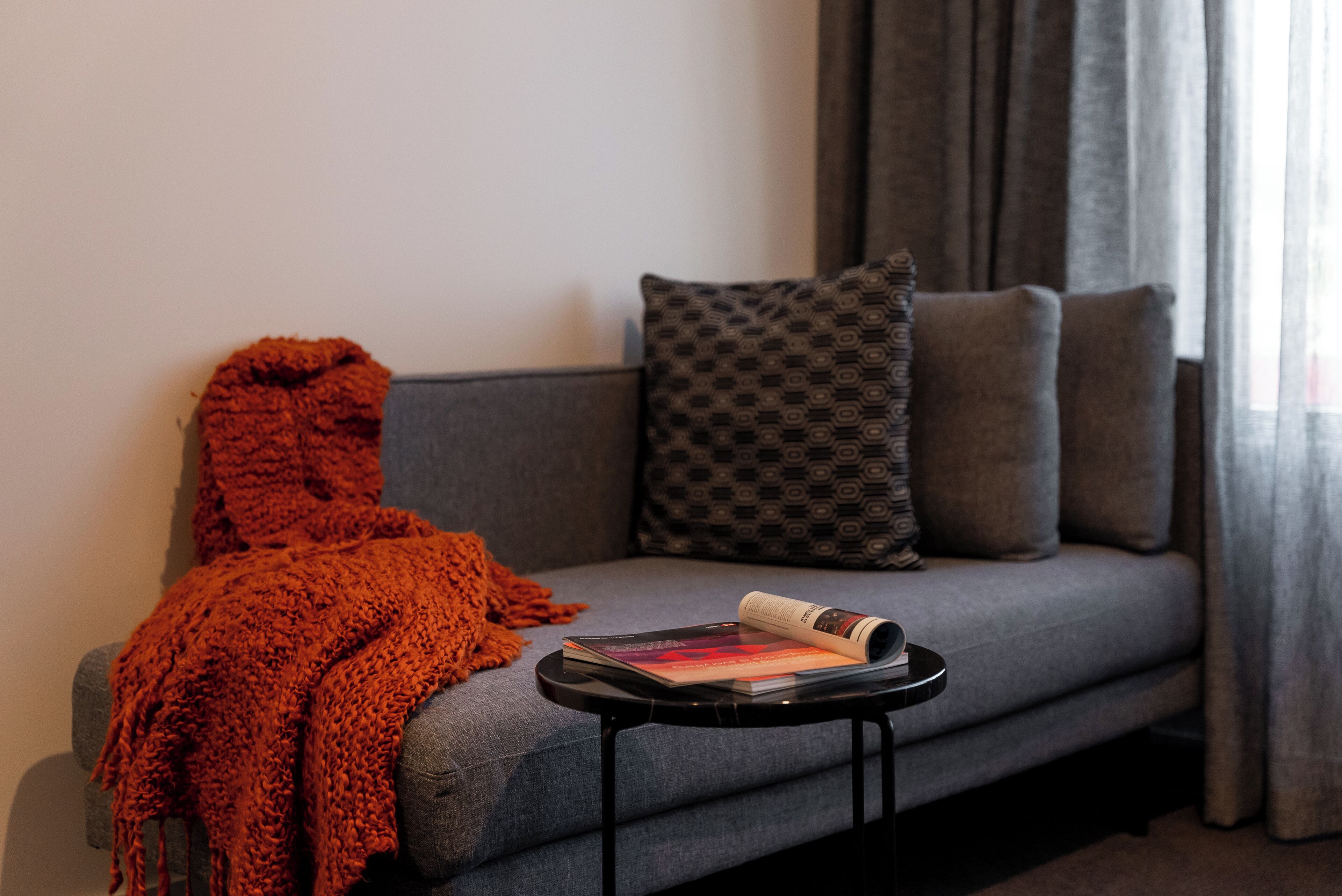 A grey sofa with patterned cushions, a knitted orange throw, and a magazine on a small black side table.