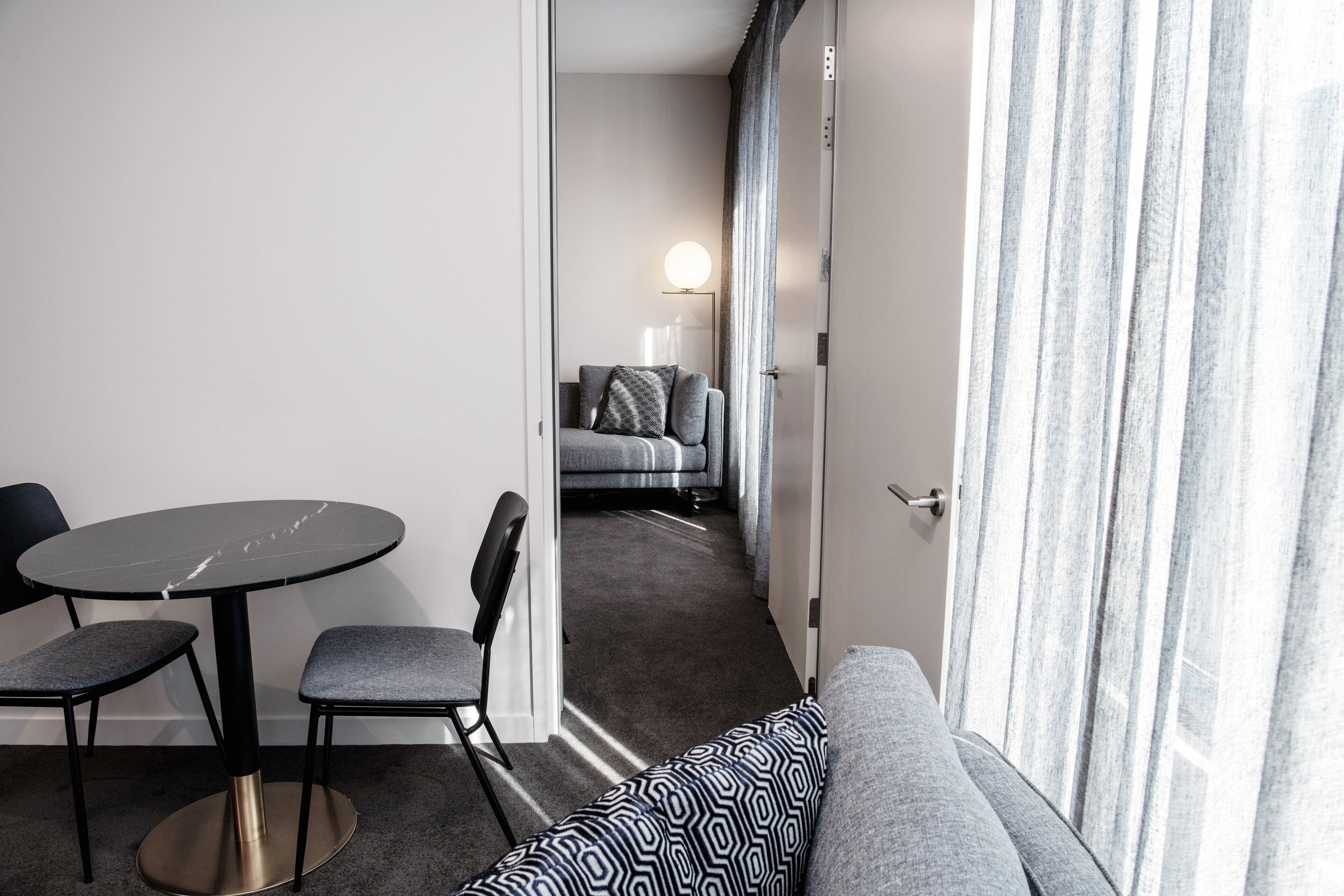 A modern hotel room with a round table, two chairs, a sofa, and a view of a balcony.