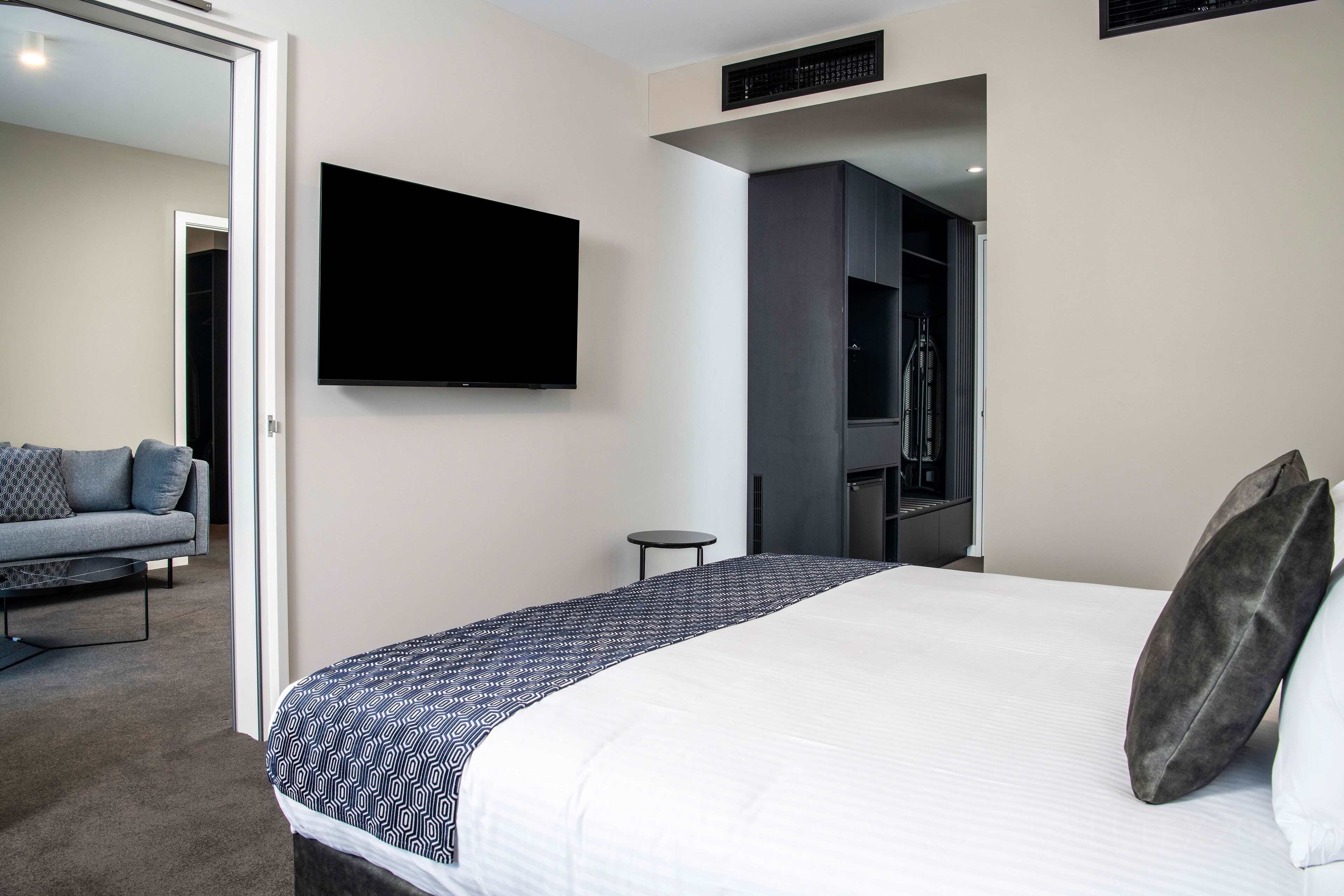 Quarto de hotel moderno com uma cama grande, uma televisão de tela plana na parede, um guarda-roupa escuro e uma pequena mesa redonda.