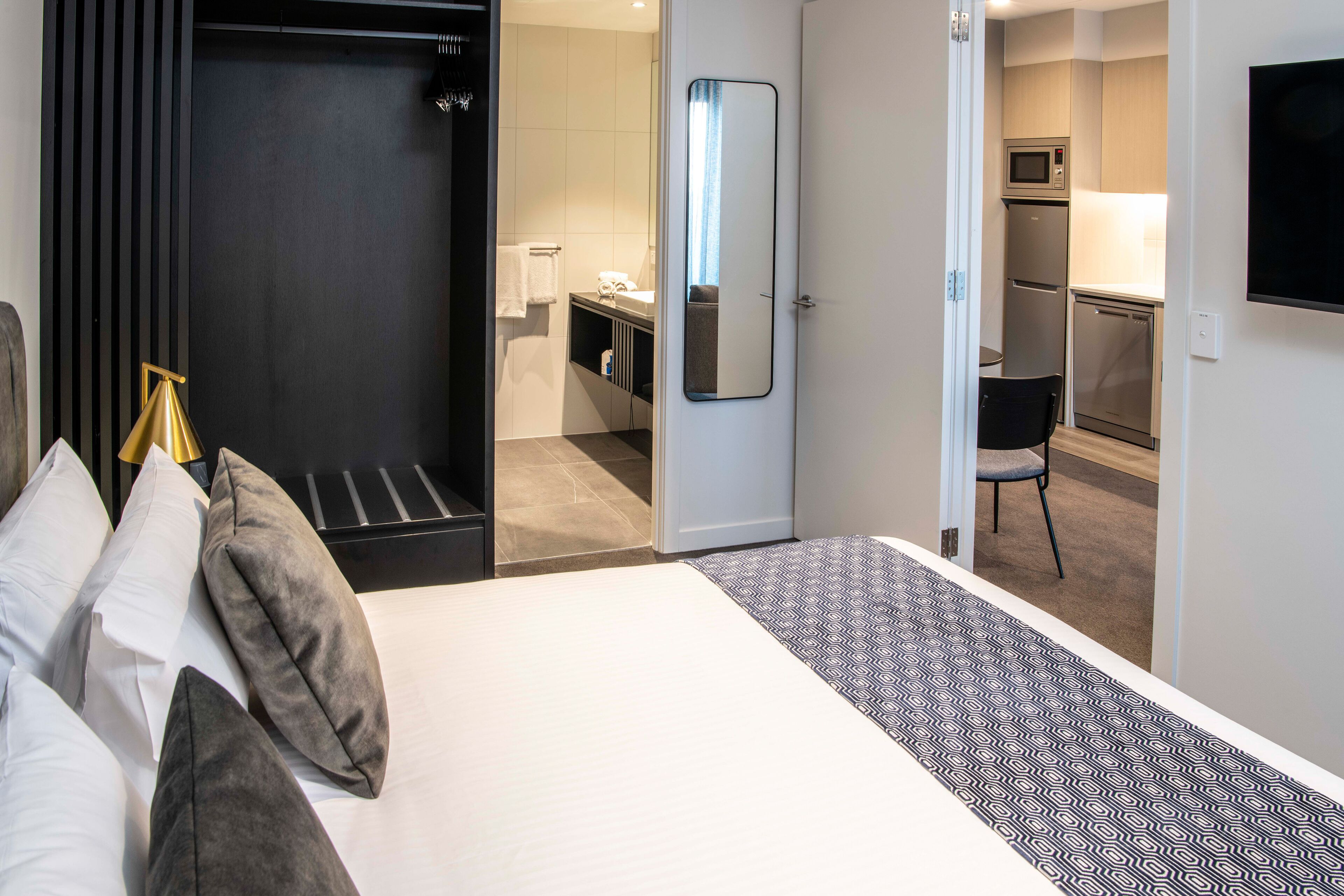 A hotel room with a bed, pillows, a wardrobe, a mirror, a desk, a chair, and a kitchenette.