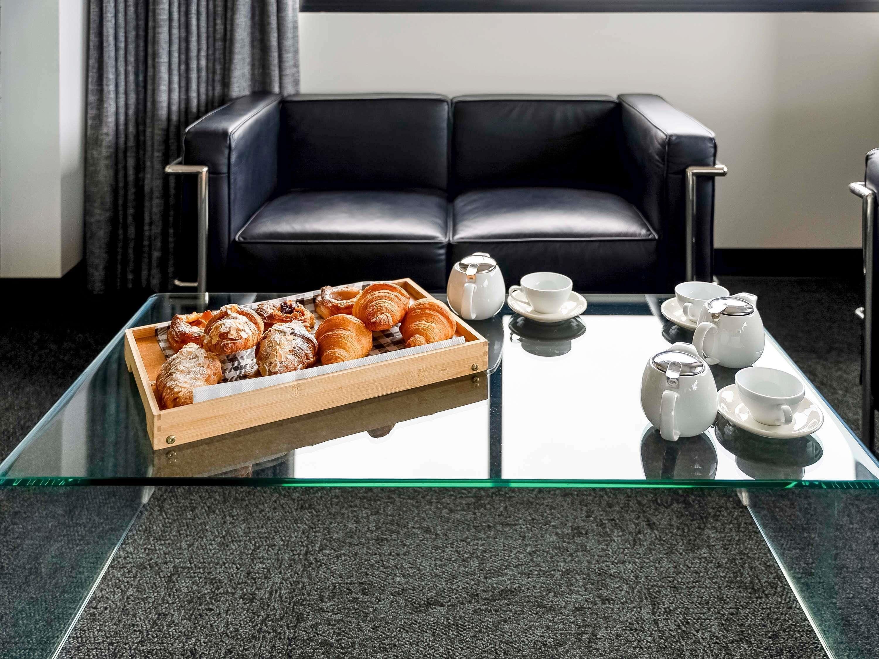 Uma mesa com tampo de vidro, com doces e jogos de chá, em um quarto moderno com sofá de couro preto.