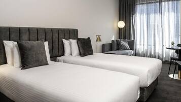 Habitación de hotel con dos camas, un escritorio, una silla, una lámpara y vistas al exterior a través de cortinas translúcidas.