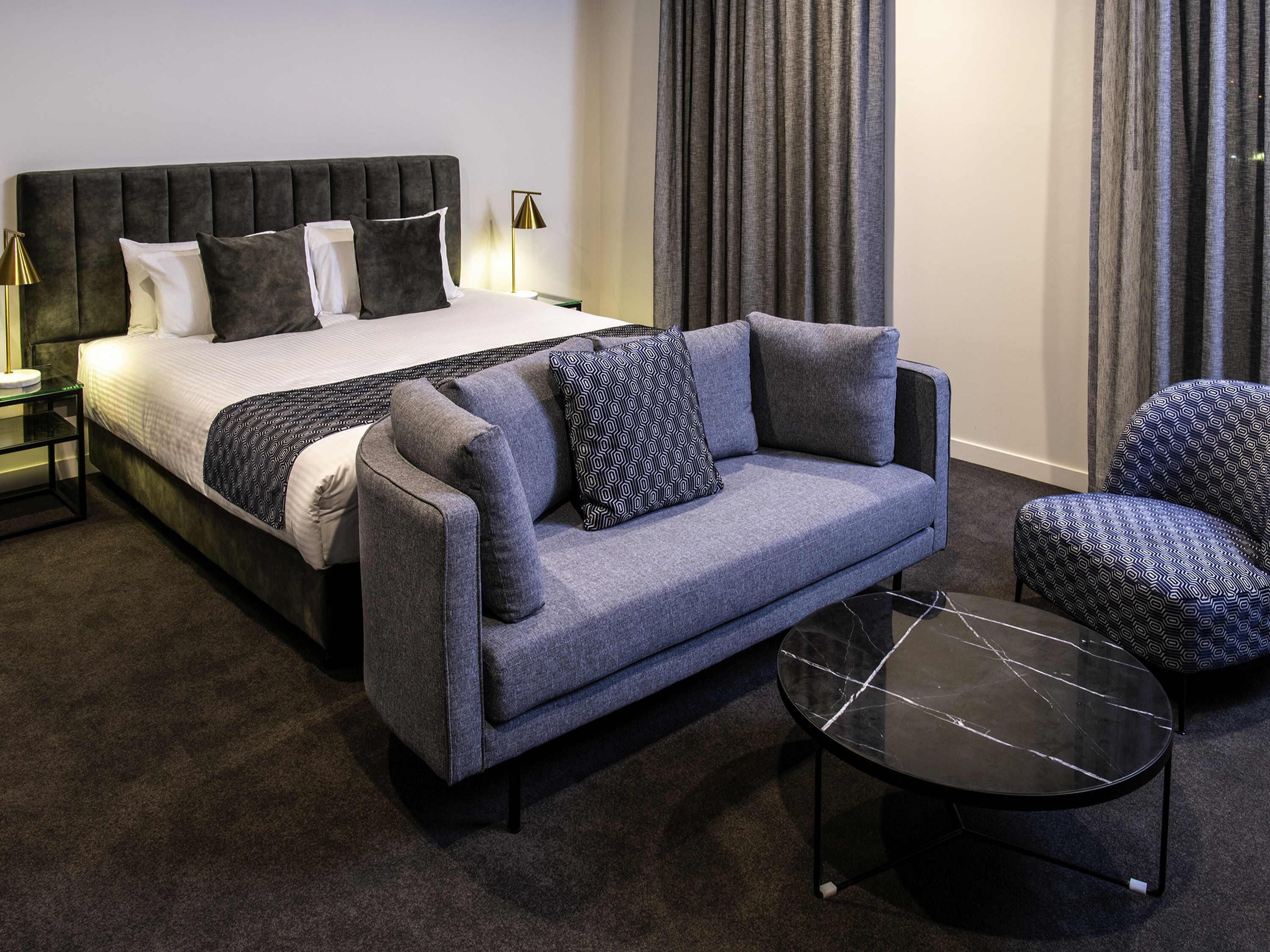 A bedroom with a bed, a grey sofa, a patterned armchair, a small black table, and a nightstand with a lamp.
