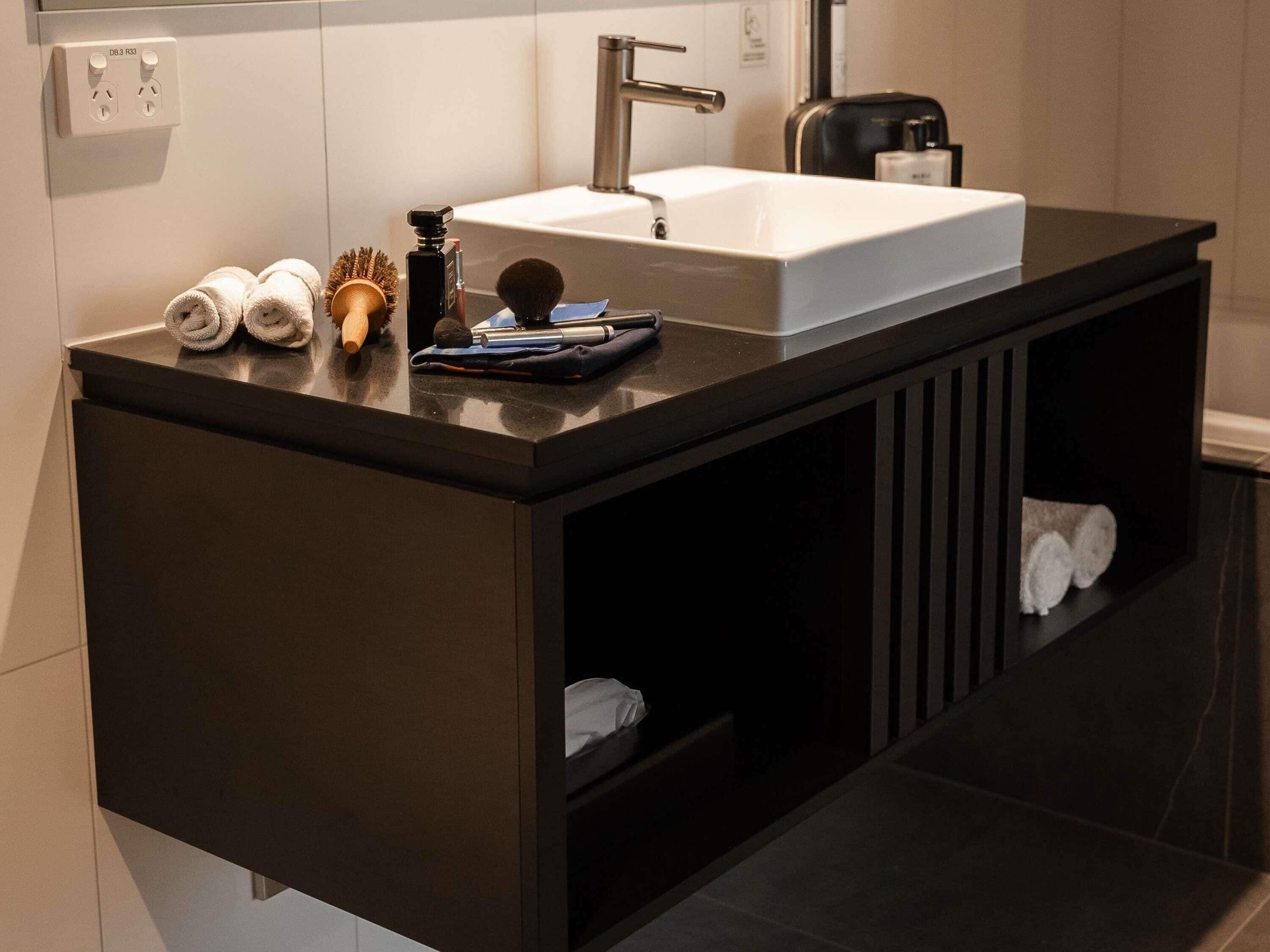 Un baño moderno con encimera oscura, un lavabo blanco y un espejo encima. Hay toallas y un cepillo de pelo en la encimera.