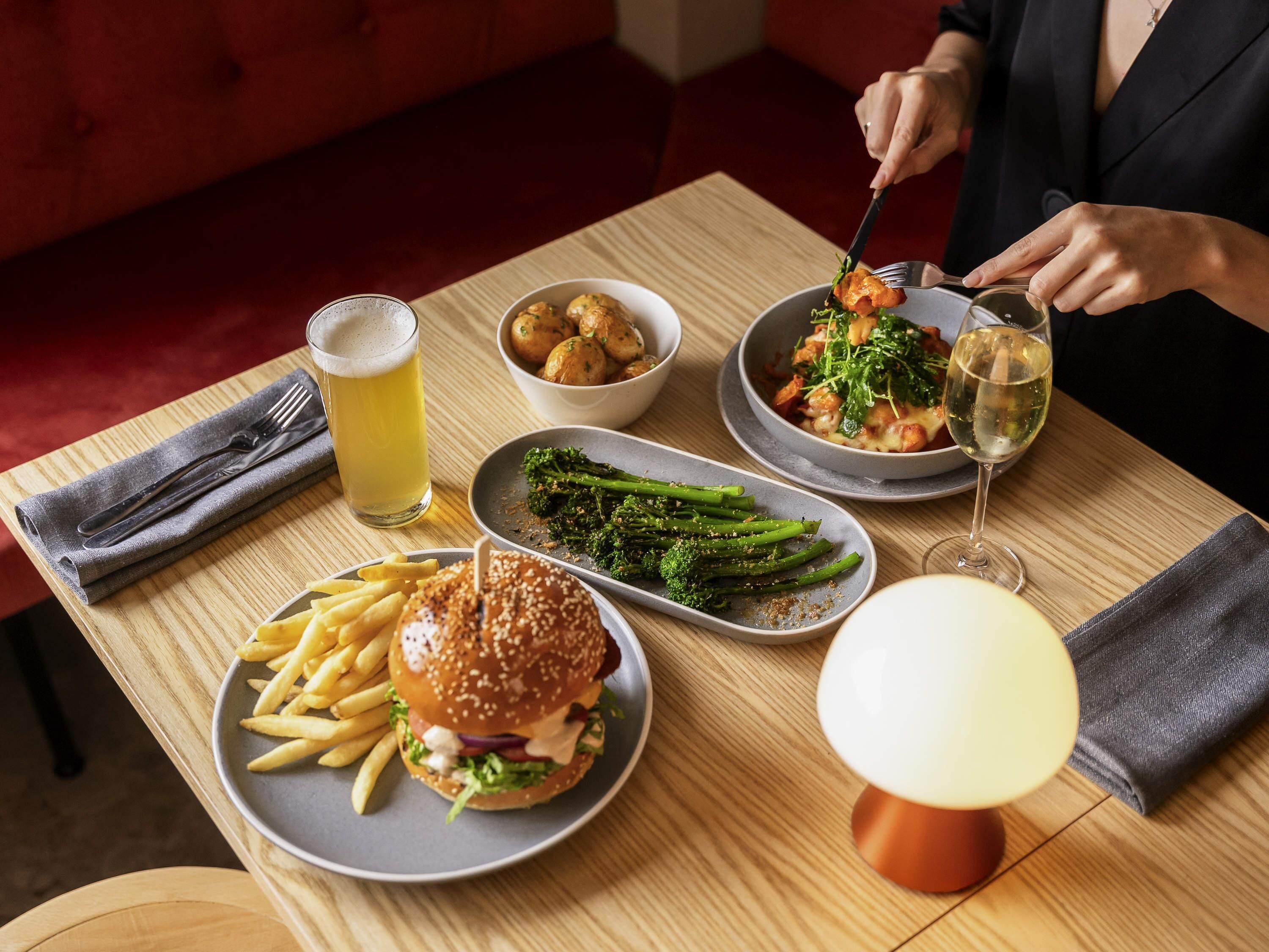 A dining table with a burger, fries, broccoli, a bowl of potatoes, and a glass of beer.