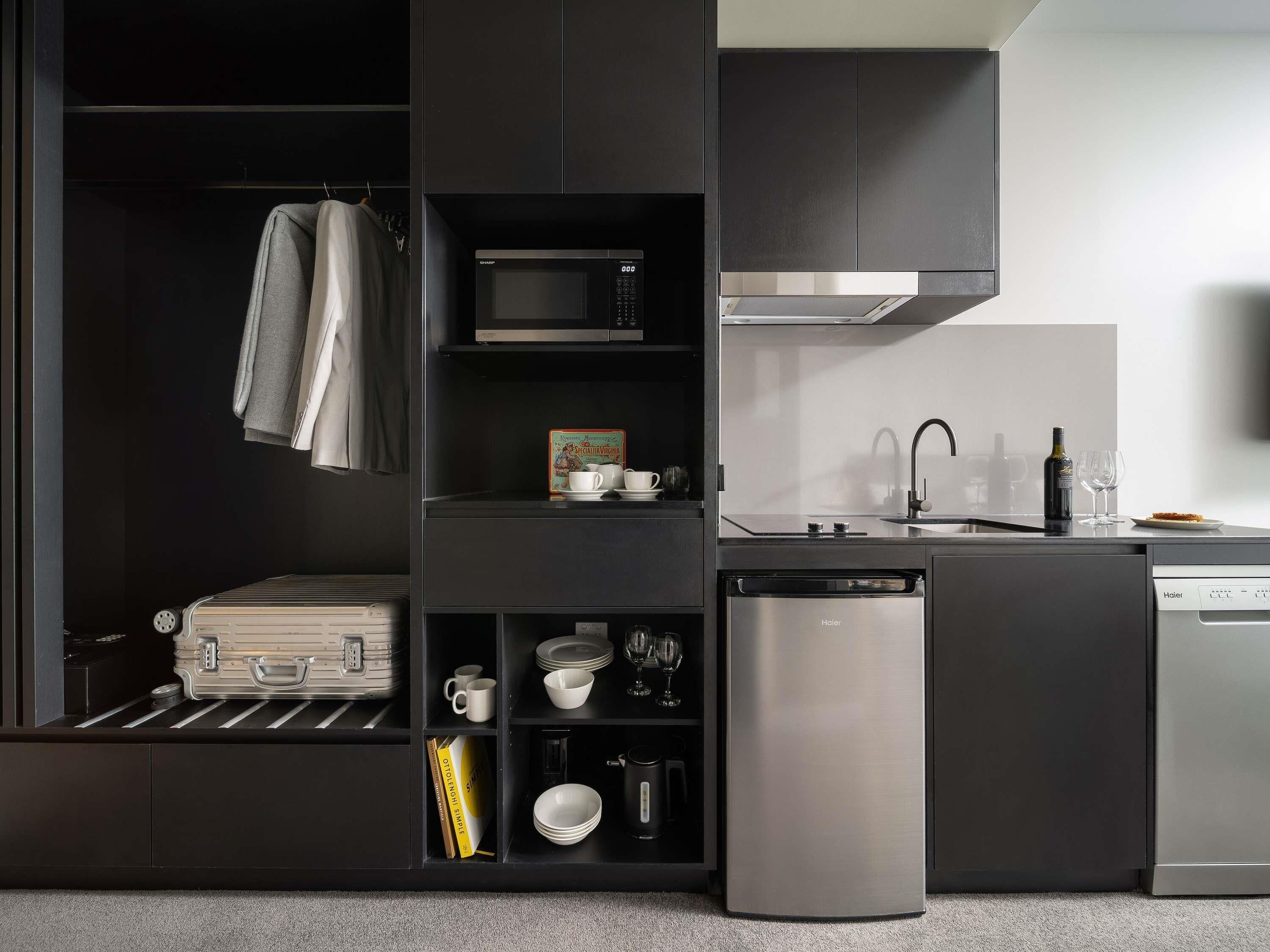 A compact kitchen with a microwave, sink, and dishwasher. There is a suitcase on the floor and a book on a shelf.