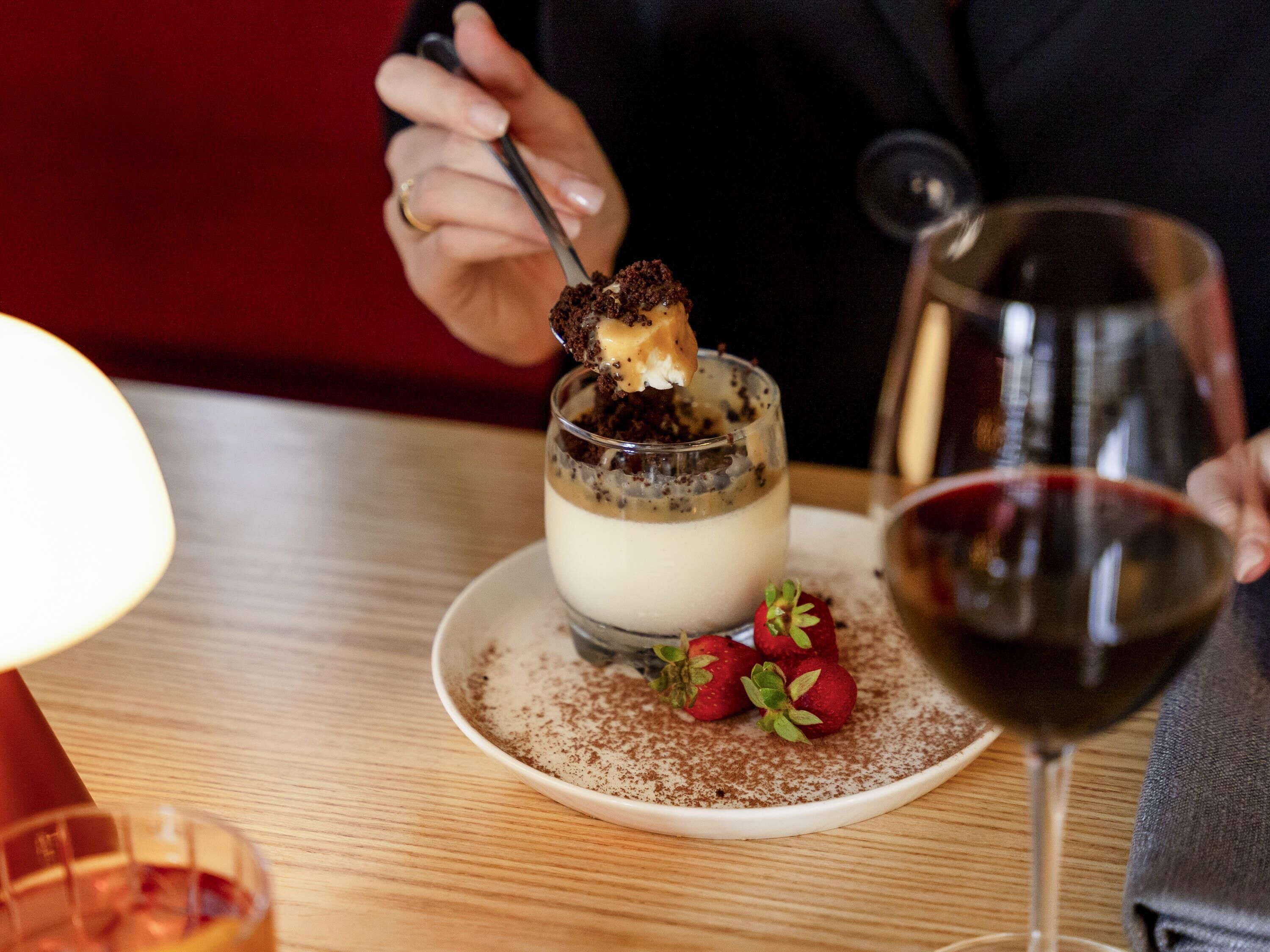 A dessert with a chocolate piece on a spoon, strawberries, and a glass of red wine on a table.
