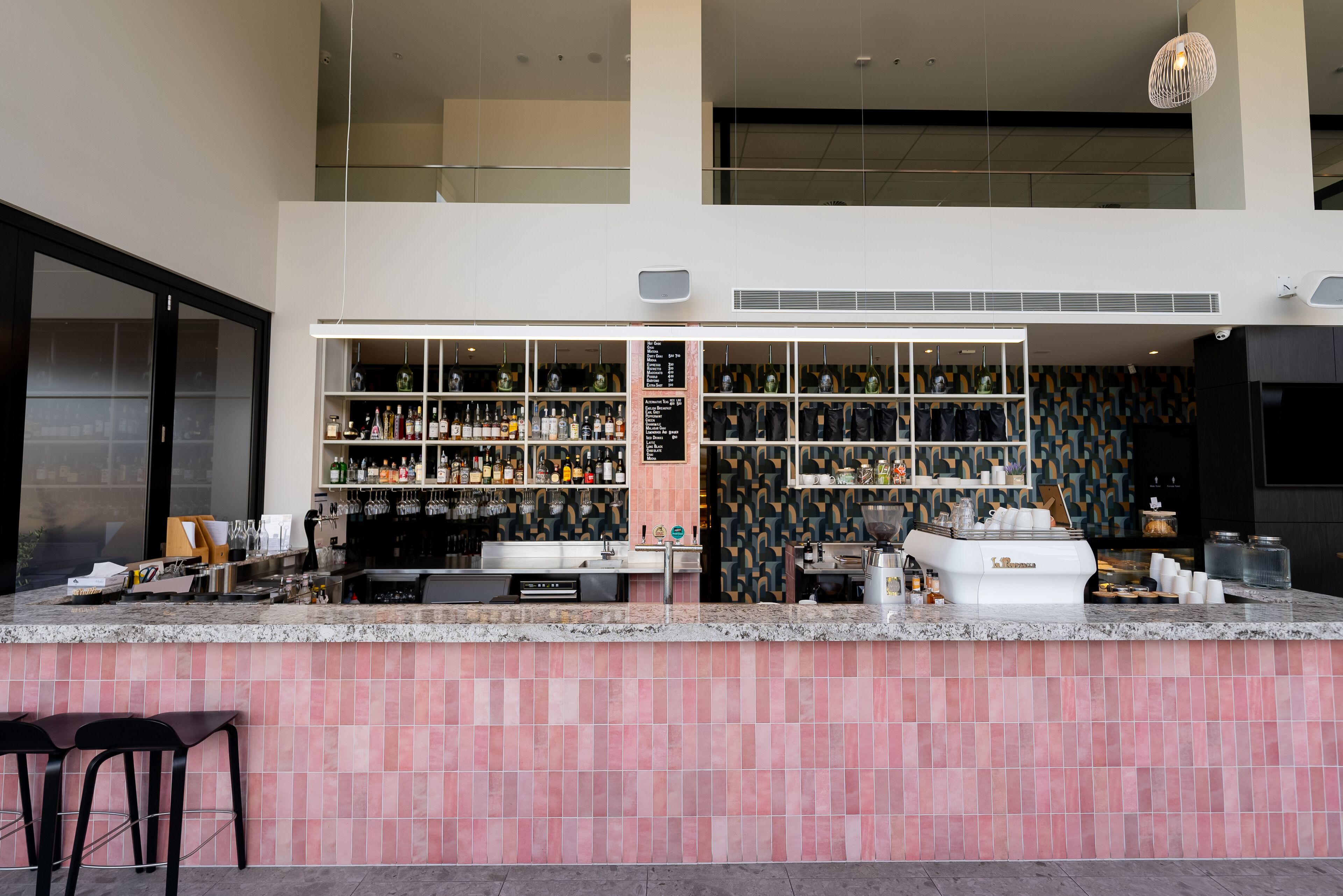 A modern bar area with a pink-tiled counter, shelves of bottles, and a coffee machine.