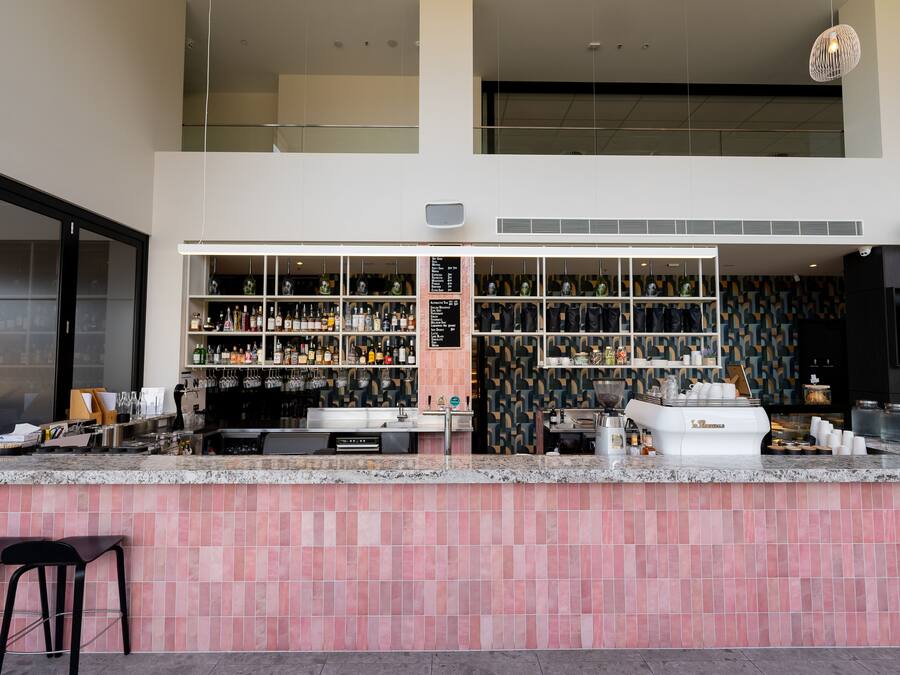 Una zona de bar moderna con mostrador de azulejos rosas, estanterías llenas de botellas y una máquina de café.