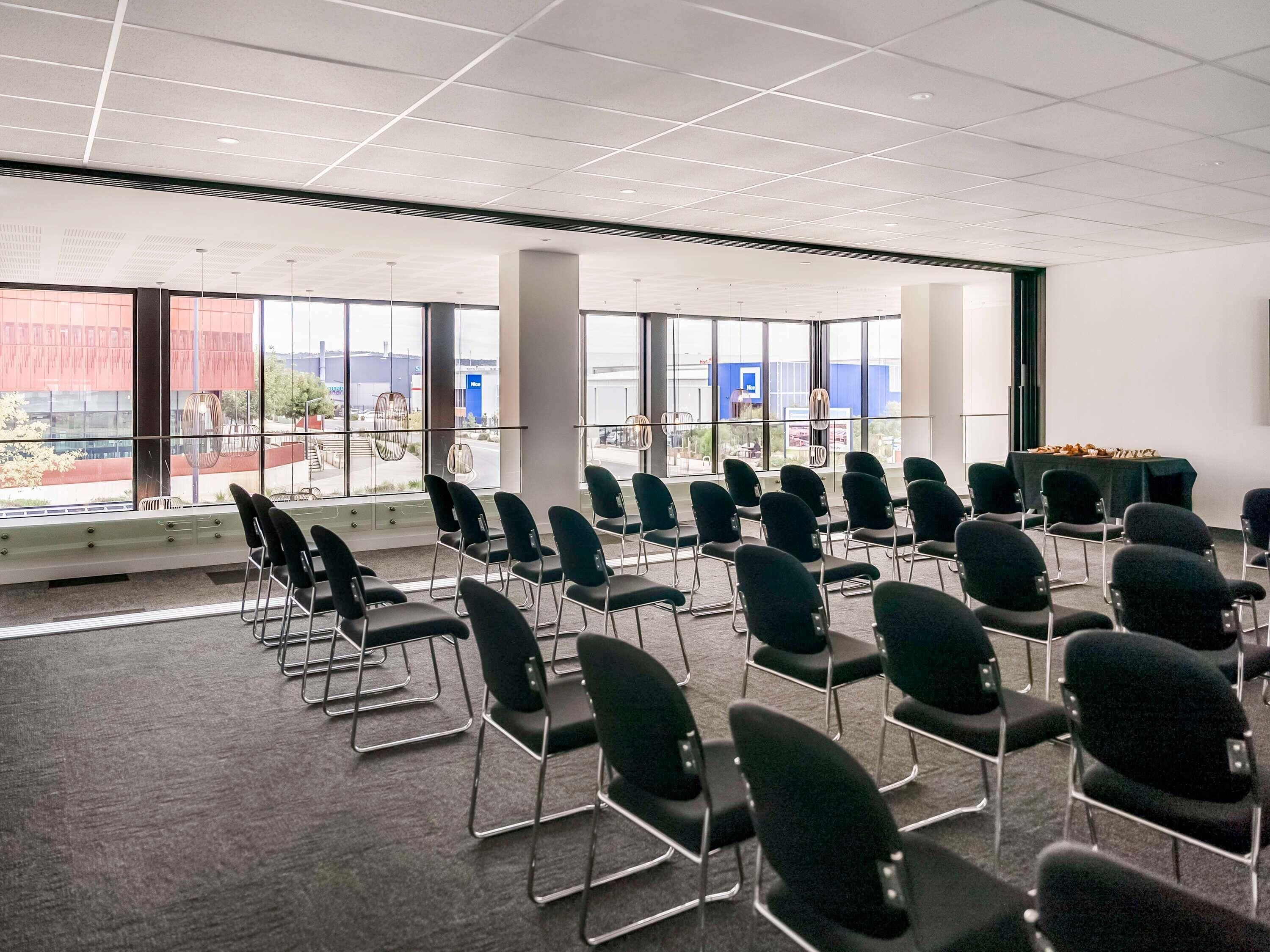 Un salón de conferencias con filas de sillas, ventanas amplias y una mesa con comida.
