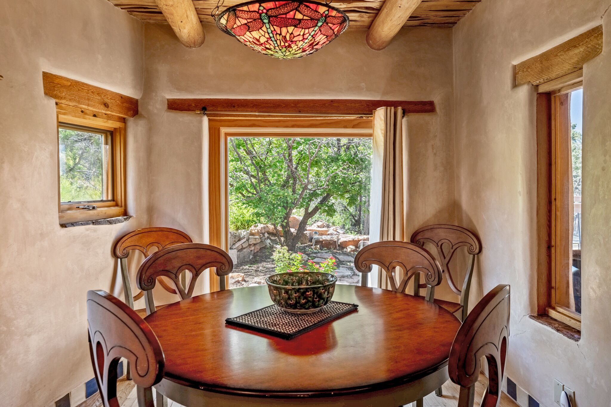 Un coin repas avec une table ronde en bois, quatre chaises, un luminaire en vitrail et une vue sur un arbre et des rochers, visible à travers une grande fenêtre.