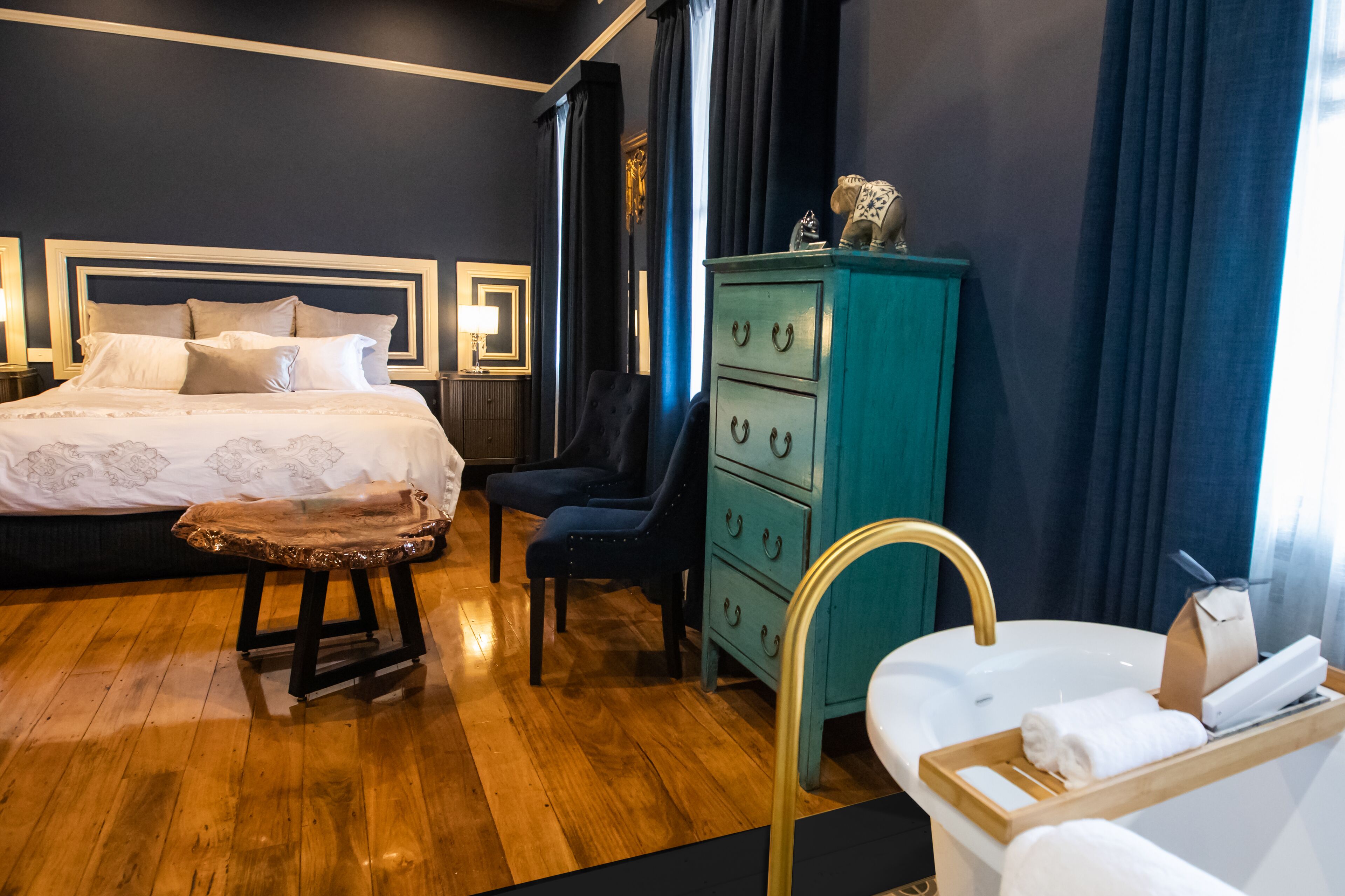 Habitación de hotel con una cama grande, un banco de madera, un aparador verde azulado, una silla y un lavabo blanco con toallas.