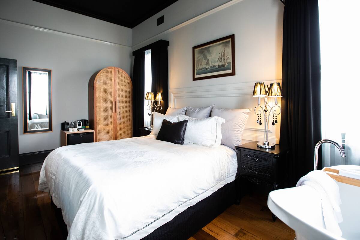 A large bed with white linens, a wooden wardrobe, a nightstand with lamps, and a painting on the wall.