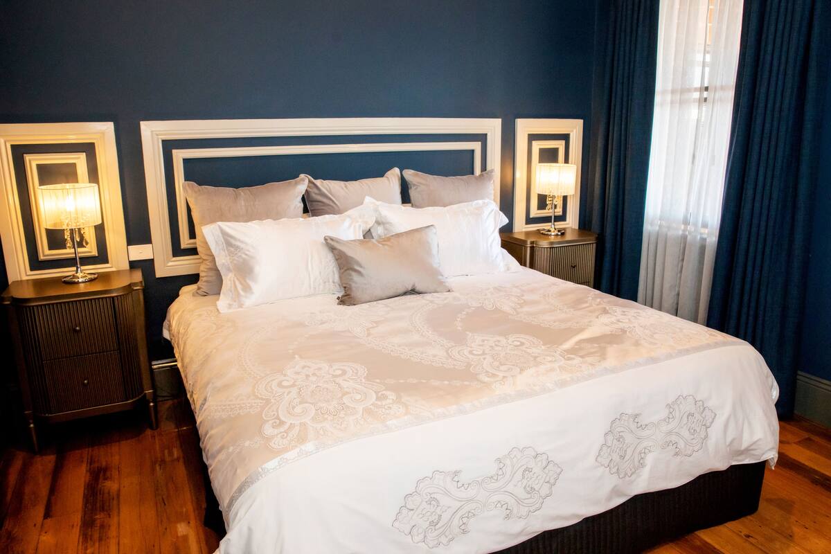 A neatly made bed with a white and beige patterned bedspread, flanked by two bedside tables with lamps, in a room with dark blue walls and wooden flooring.