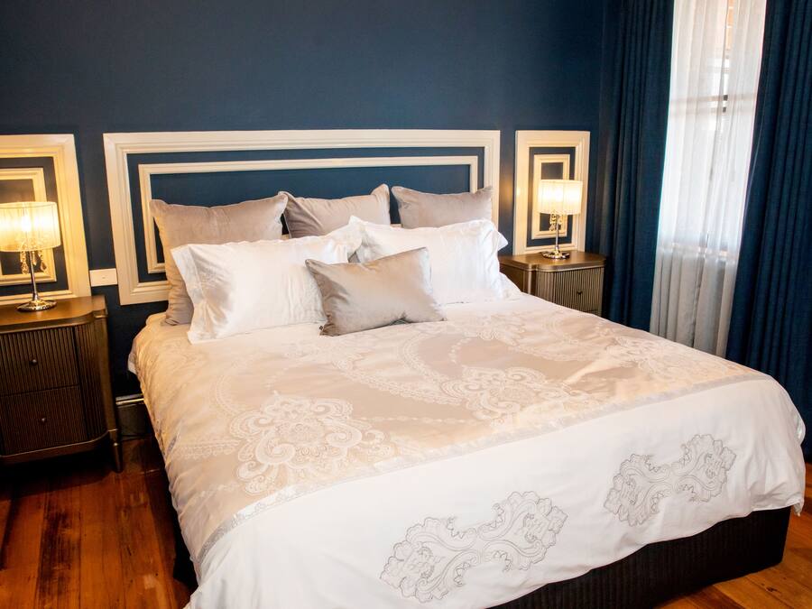 Una cama bien tendida con una colcha blanca y color crema estampada, flanqueada por dos mesitas de noche con lámparas, en una habitación con paredes azul oscuro y suelo de madera.