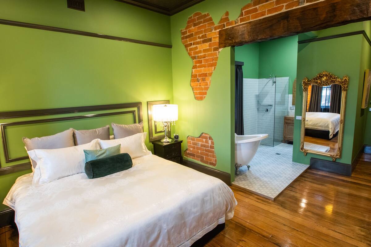 A bedroom with a large bed, a nightstand with a lamp, a brick wall mural, a bathroom with a bathtub, and a mirror with an ornate frame.