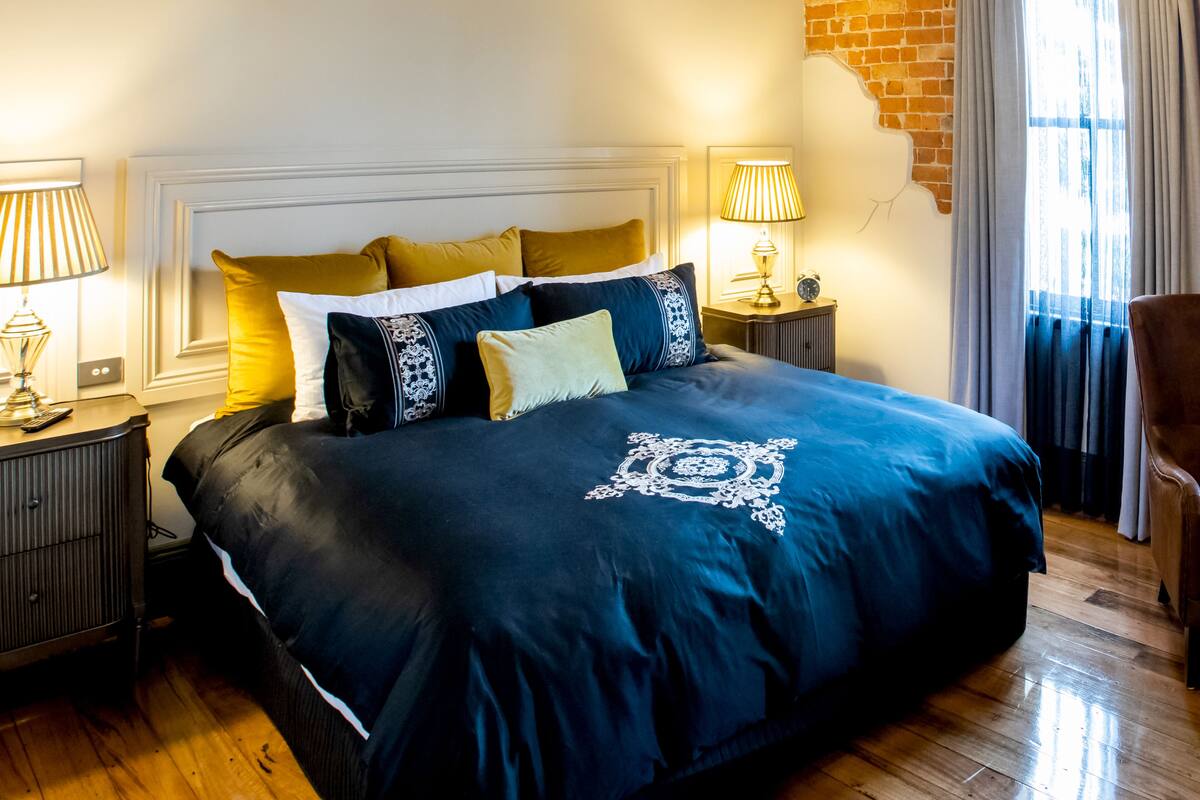 A neatly made bed with a dark blue bedspread and decorative pillows, two bedside lamps, a wooden nightstand, and a brick wall.