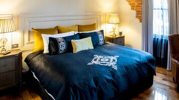 Una cama bien tendida con una colcha azul oscuro y almohadones decorativos, dos lámparas de noche, una mesita de madera y una pared de ladrillo.