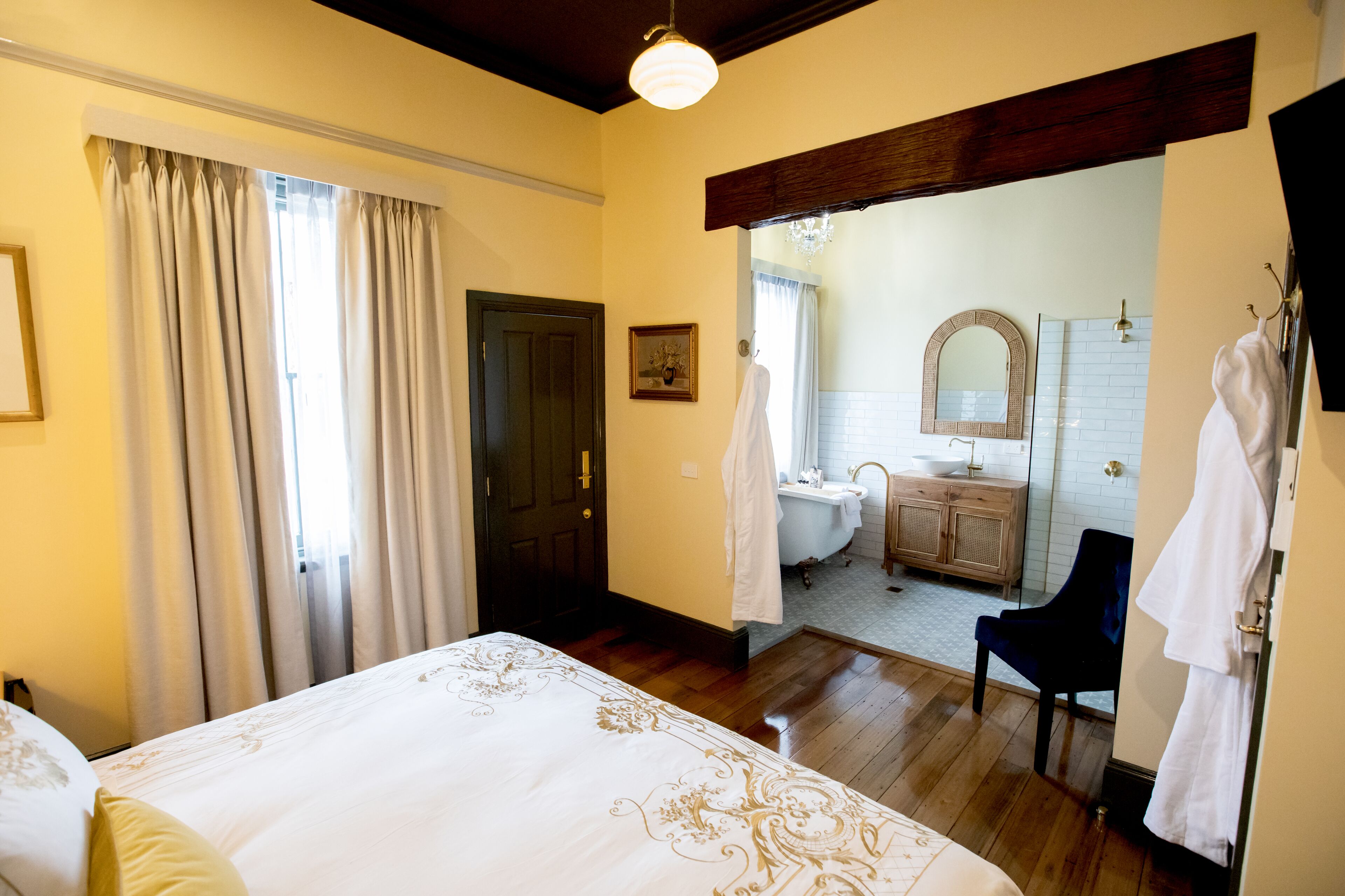 Une chambre avec un grand lit, un plancher en bois, une porte et une vue sur une salle de bain équipée d’une baignoire et d’un miroir.