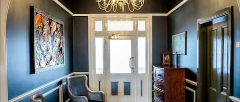 A hallway with a chandelier, two armchairs, a round rug, and a wooden cabinet.