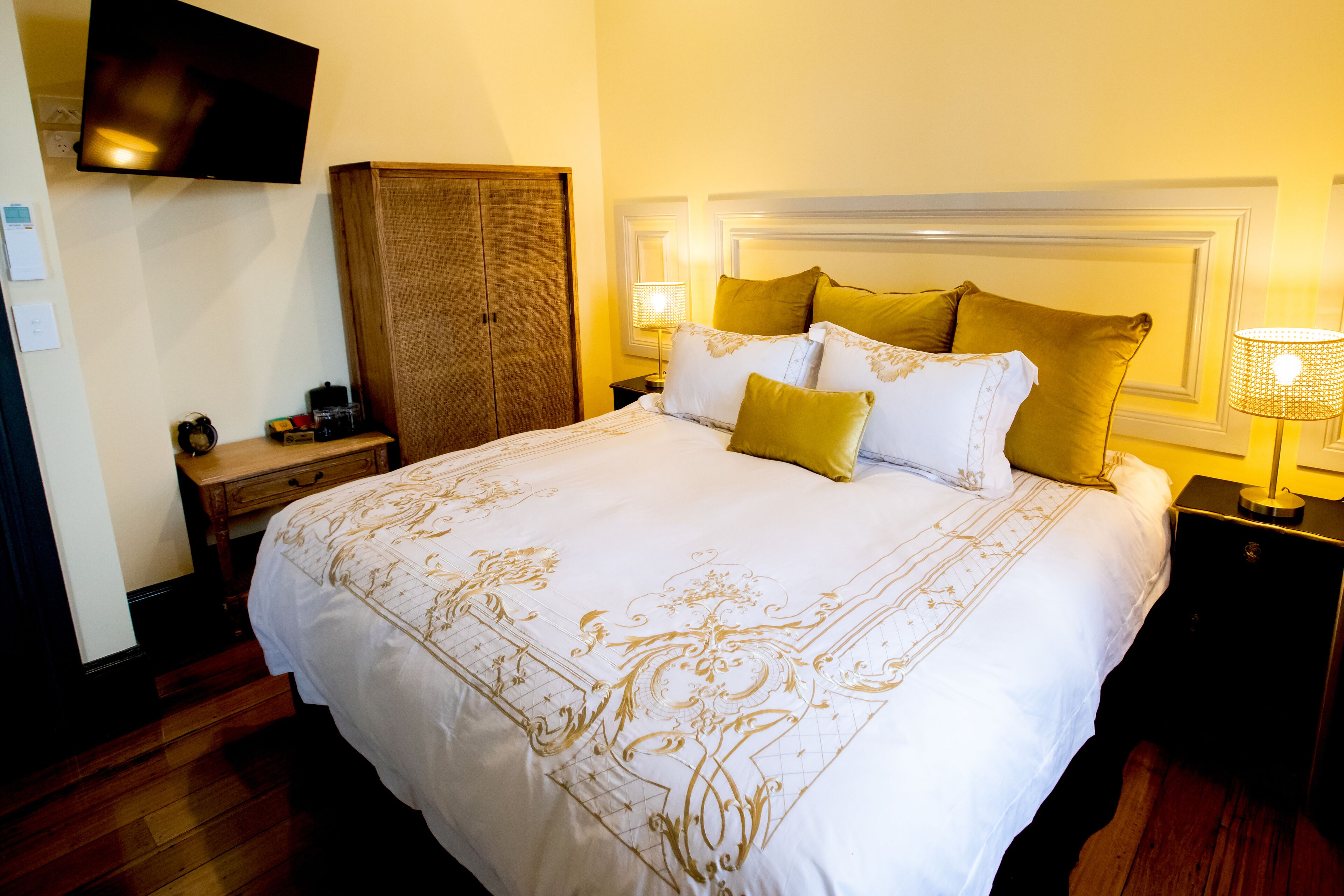 A large bed with a white and gold bedspread, two nightstands with lamps, a wooden wardrobe, and a flat-screen TV mounted on the wall.
